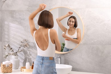 Smiling woman applying roll-on deodorant near mirror at home, back view