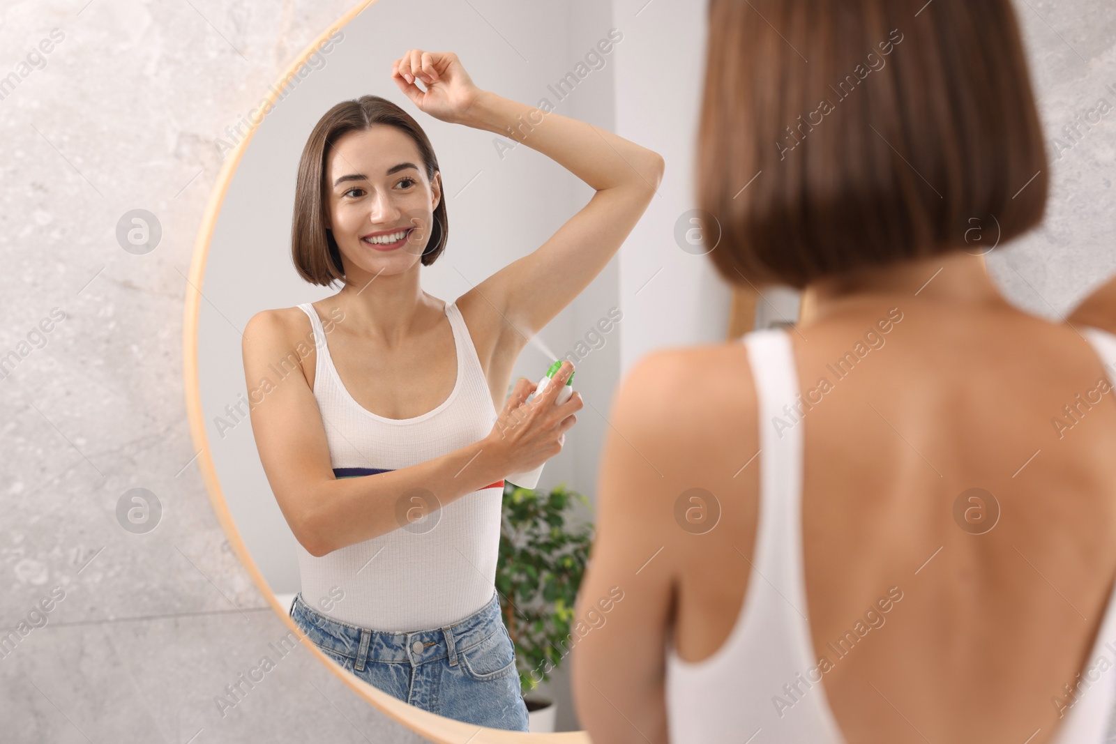 Photo of Smiling woman applying spray deodorant near mirror at home, back view