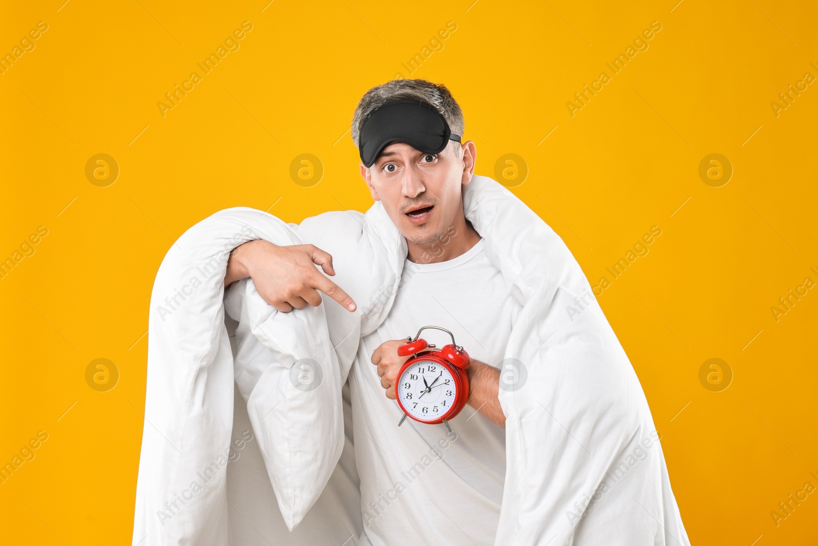 Photo of Overslept man with sleep mask and alarm clock wrapped in blanket on orange background