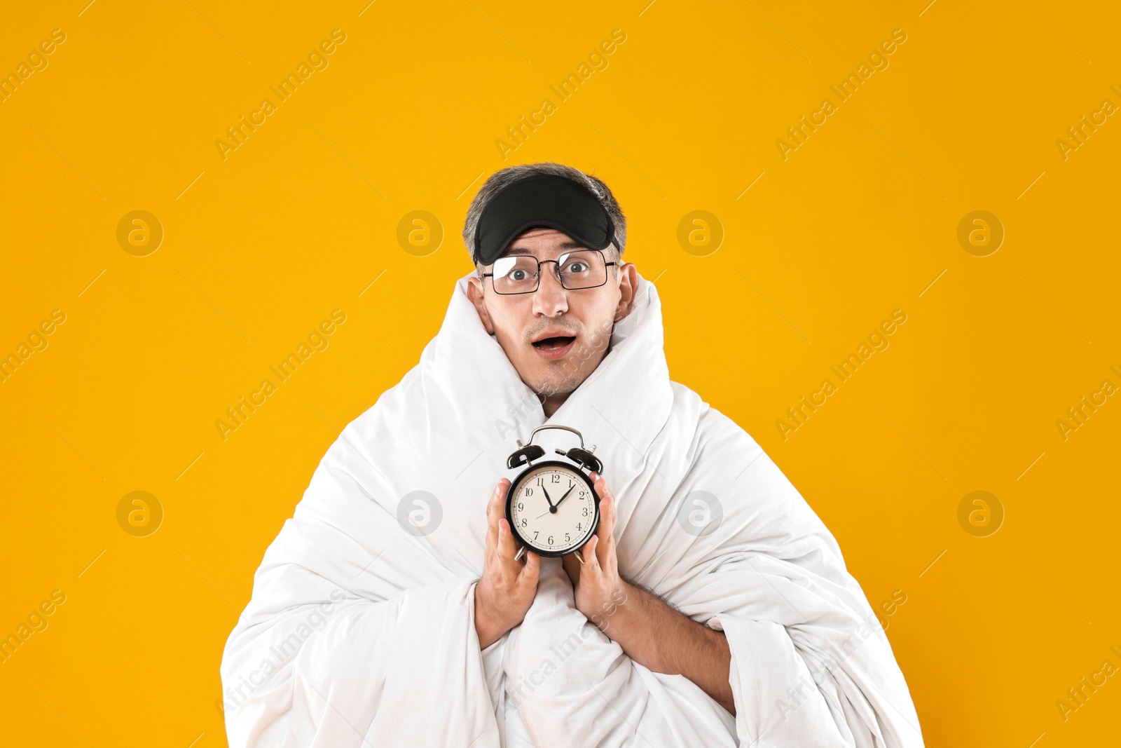 Photo of Overslept man with sleep mask and alarm clock wrapped in blanket on orange background