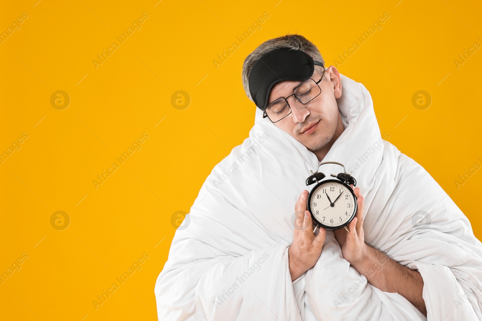 Photo of Overslept man with sleep mask and alarm clock wrapped in blanket on orange background, space for text