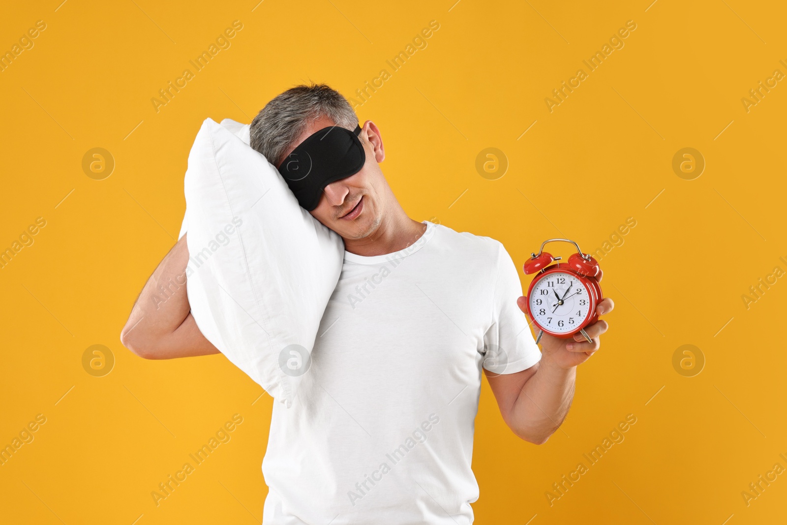 Photo of Overslept man with sleep mask, pillow and alarm clock on orange background