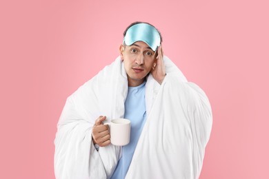 Photo of Overslept man with sleep mask and cup wrapped in blanket on pink background