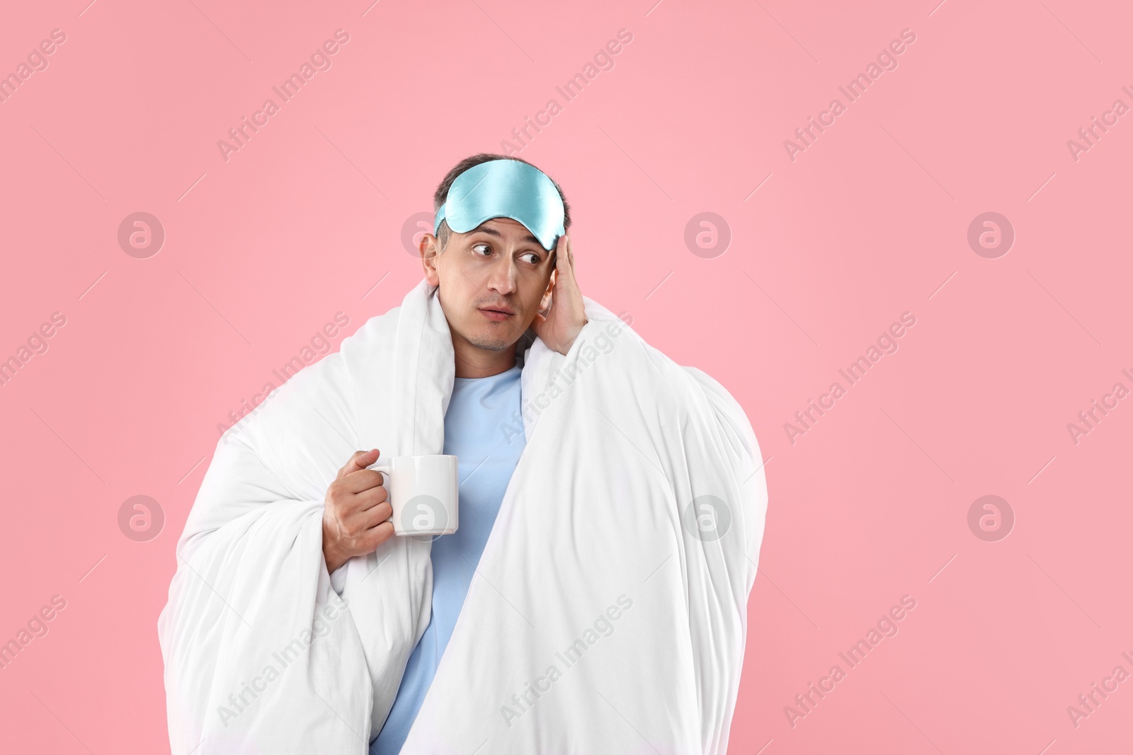 Photo of Overslept man with sleep mask and cup wrapped in blanket on pink background, space for text