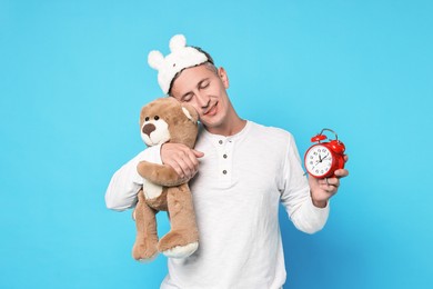 Photo of Overslept man with sleep mask, teddy bear and alarm clock on light blue background