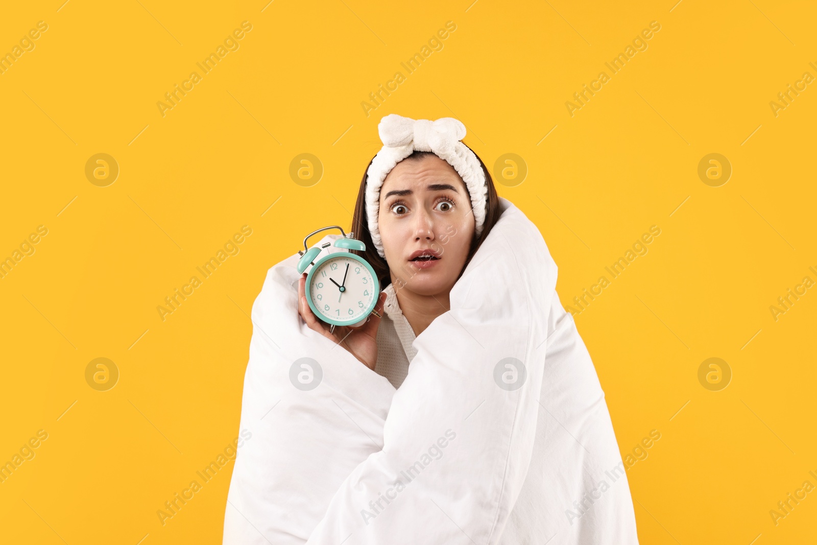 Photo of Overslept young woman with alarm clock wrapped in blanket on yellow background