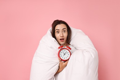 Overslept young woman with alarm clock wrapped in blanket on pink background