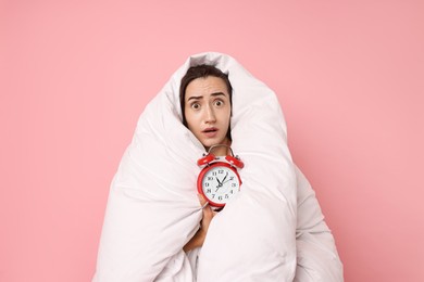 Overslept young woman with alarm clock wrapped in blanket on pink background