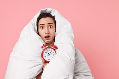 Overslept young woman with alarm clock wrapped in blanket on pink background