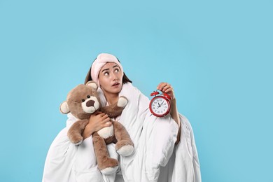 Overslept young woman with teddy bear and alarm clock wrapped in blanket on light blue background