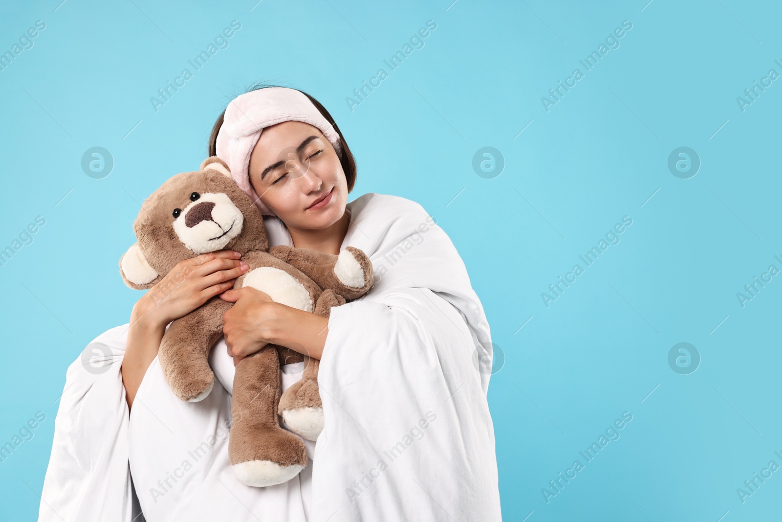 Photo of Overslept young woman with teddy bear wrapped in blanket on light blue background. Space for text