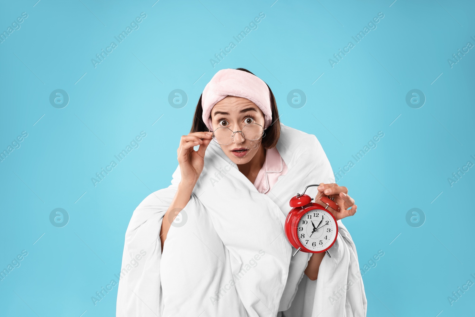 Photo of Overslept young woman with glasses and alarm clock wrapped in blanket on light blue background