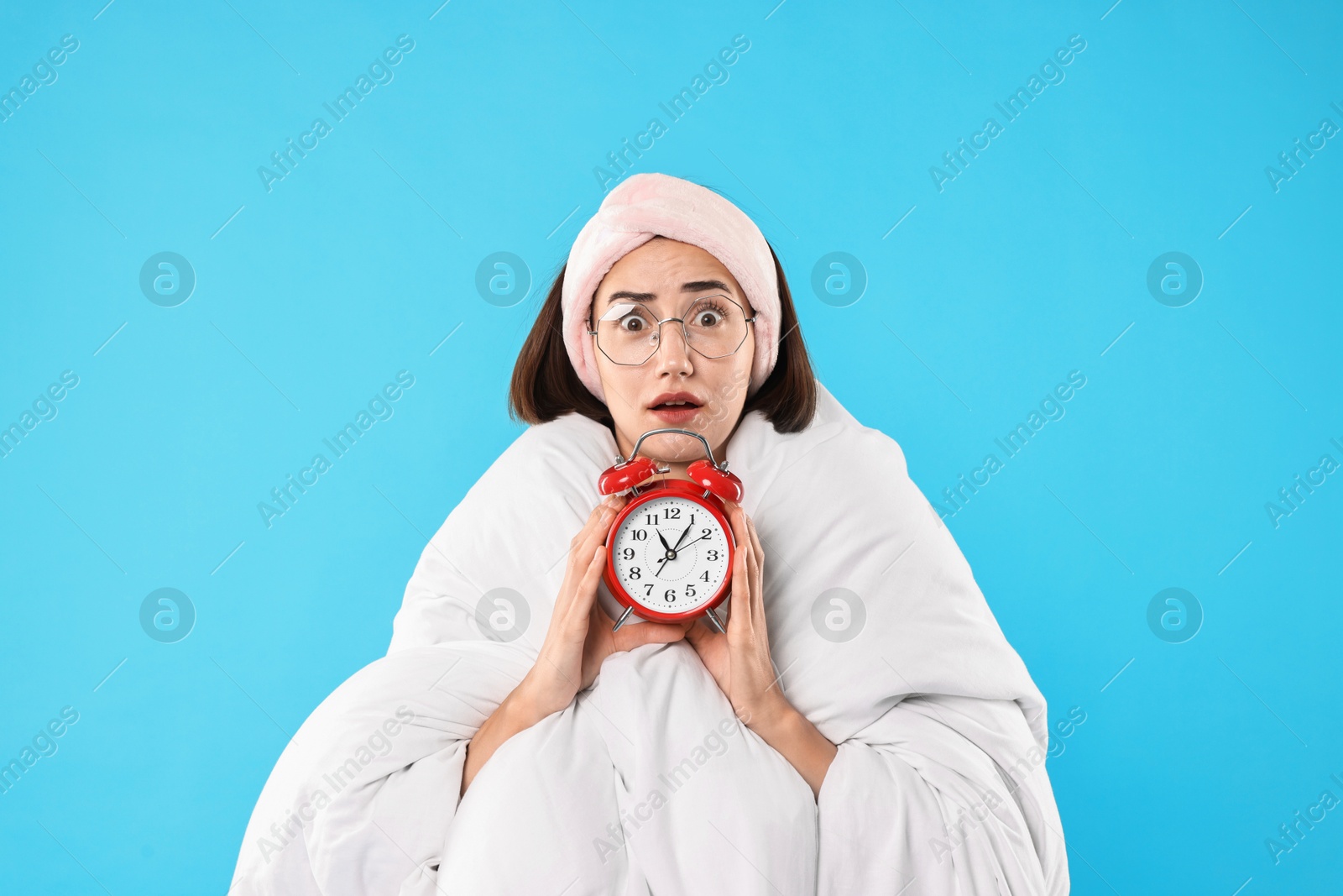 Photo of Overslept young woman with glasses and alarm clock wrapped in blanket on light blue background