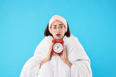 Overslept young woman with glasses and alarm clock wrapped in blanket on light blue background