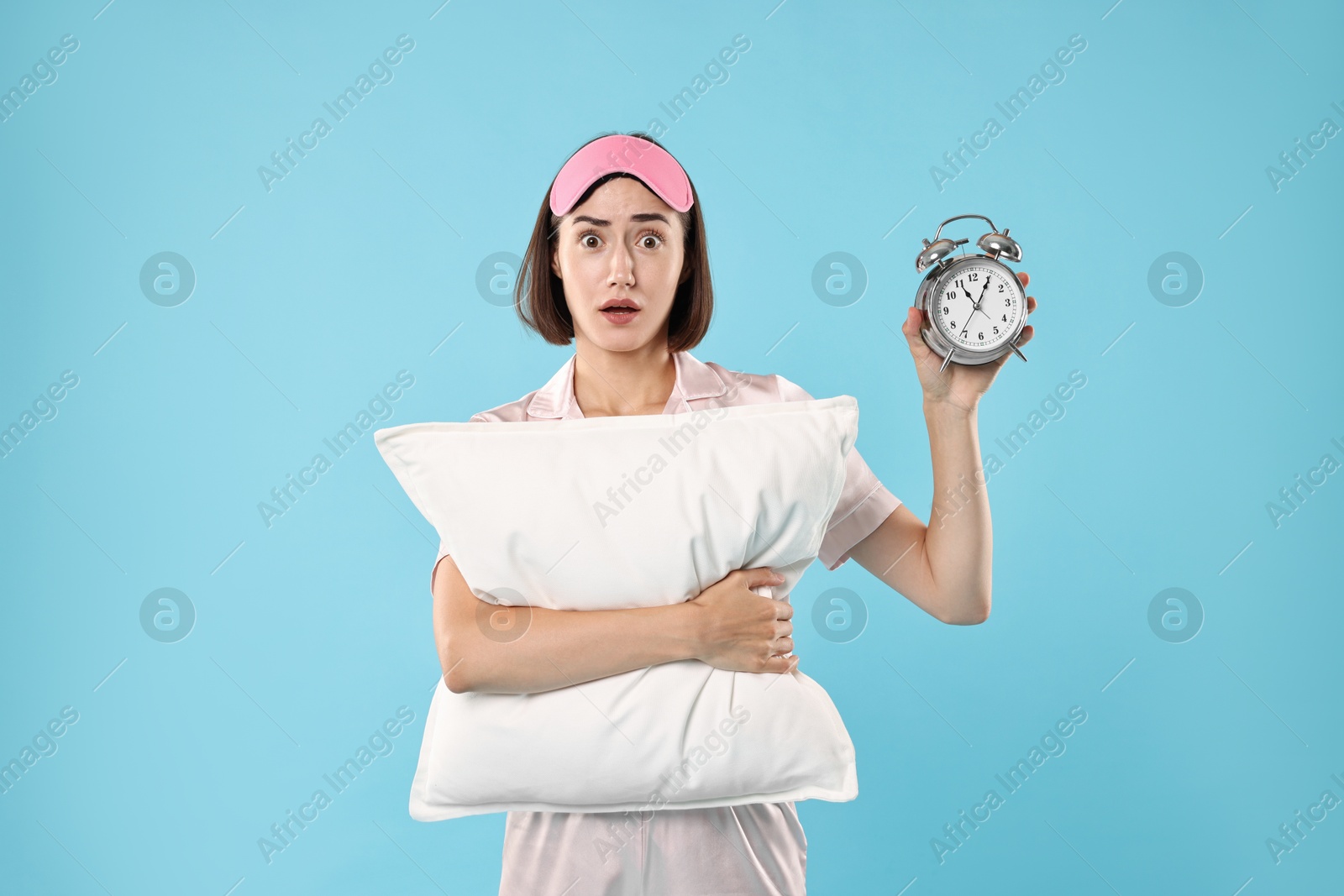 Photo of Overslept young woman with sleep mask, pillow and alarm clock on light blue background