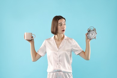 Overslept young woman with cup and alarm clock on light blue background