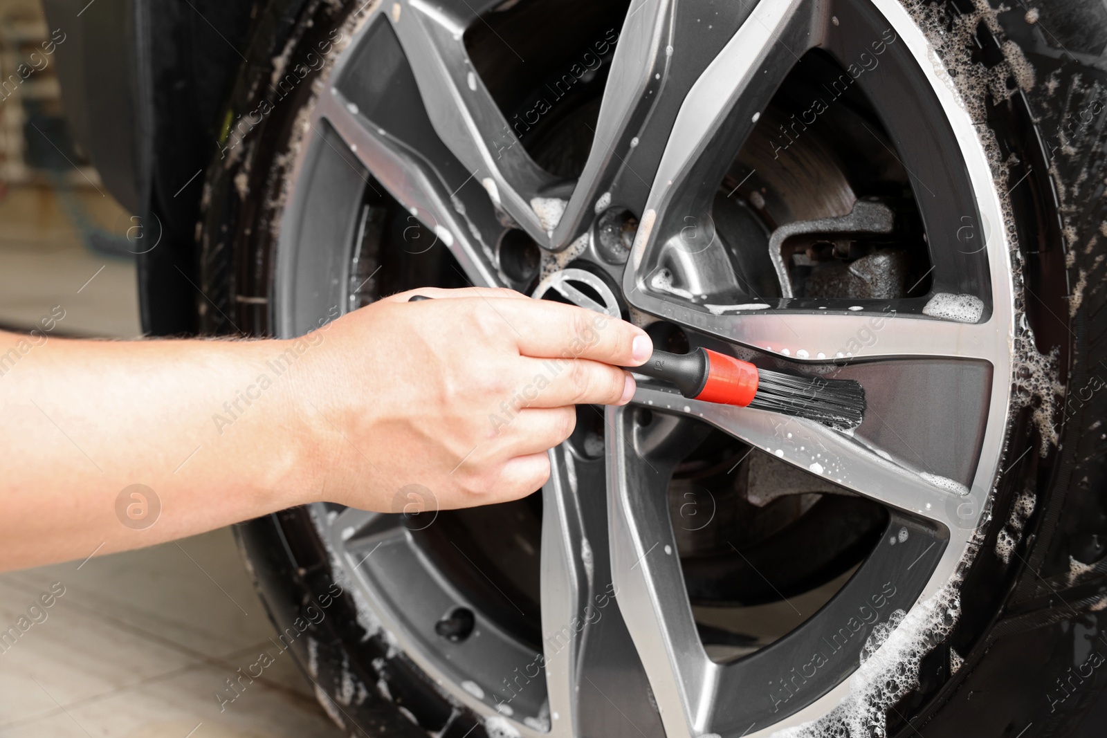 Photo of Man cleaning car wheel with brush indoors, closeup