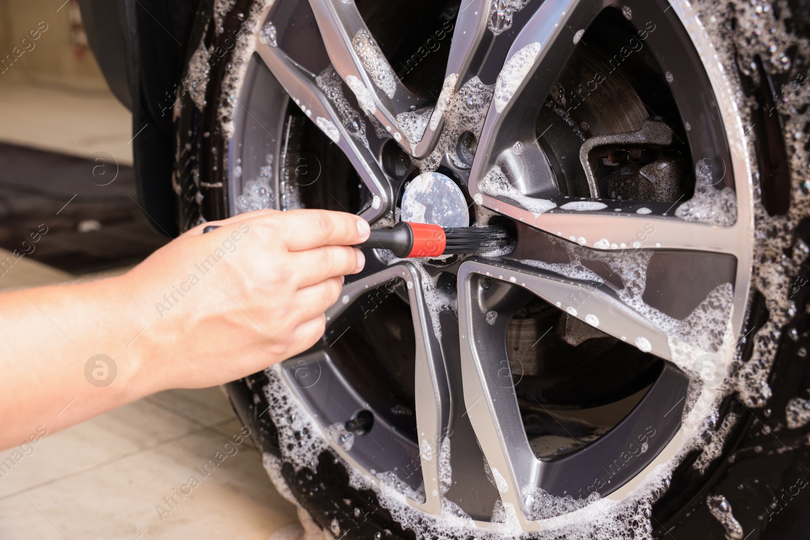 Photo of Man cleaning car wheel with brush indoors, closeup