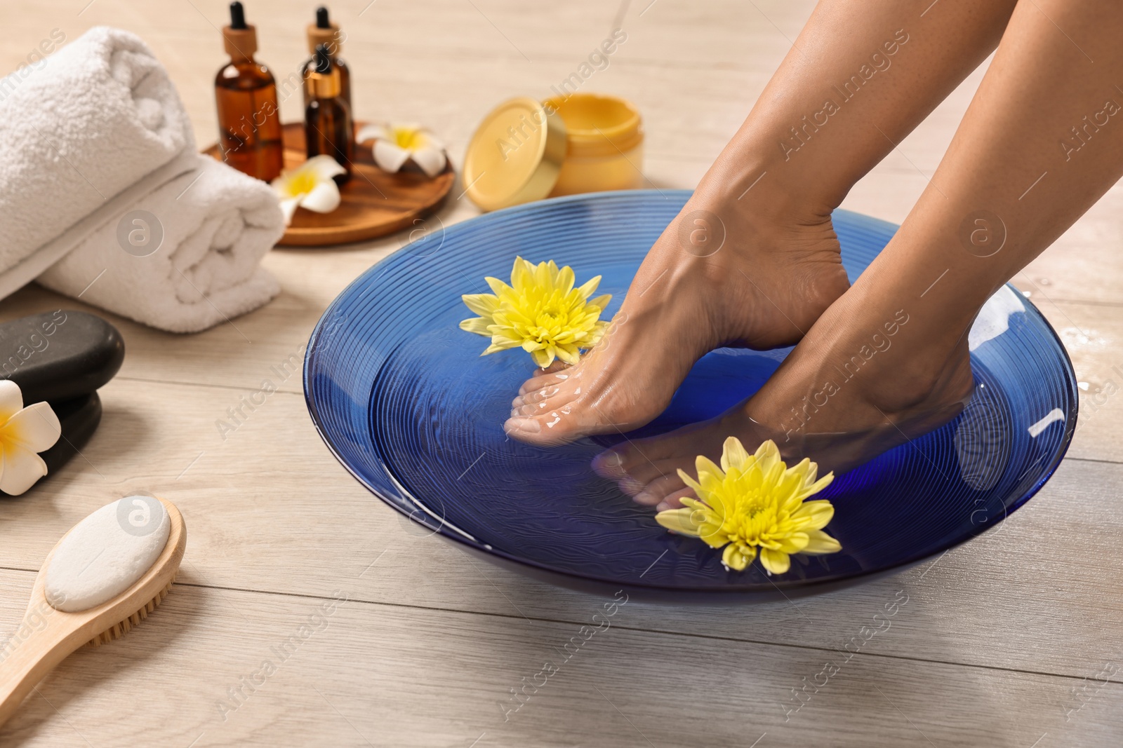 Photo of Woman soaking her feet in bowl with water and flowers on floor, closeup. Body care