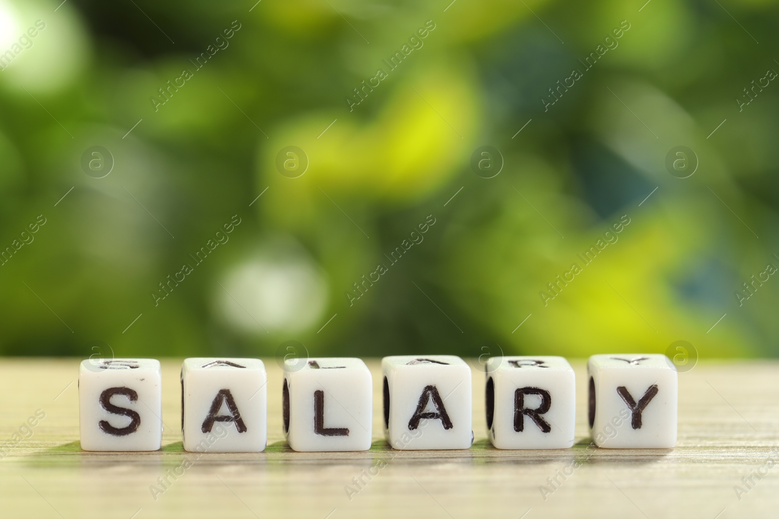 Photo of Word Salary made of cubes on light wooden table against blurred green background, closeup. Space for text