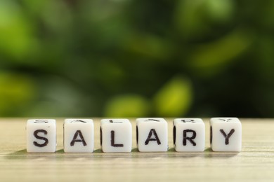 Photo of Word Salary made of cubes on light wooden table against blurred green background, closeup. Space for text