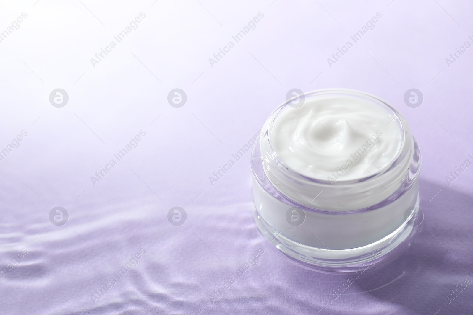 Photo of Jar with face cream in water on light violet background, space for text