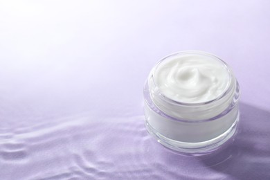 Photo of Jar with face cream in water on light violet background, space for text