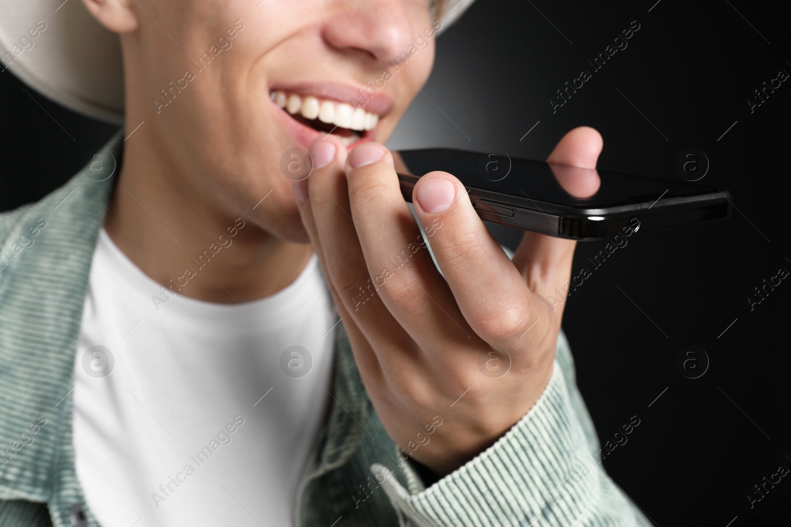 Photo of Young man recording voice message via smartphone on dark background, closeup