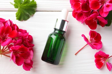 Photo of Bottle of geranium essential oil and beautiful flowers on white wooden table, flat lay