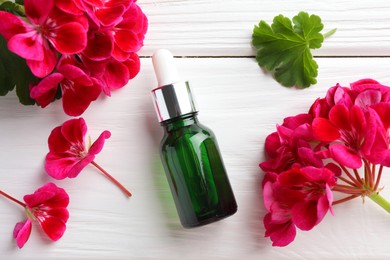 Photo of Bottle of geranium essential oil and beautiful flowers on white wooden table, flat lay