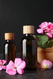 Photo of Bottles of geranium essential oil and beautiful flowers on black table, closeup