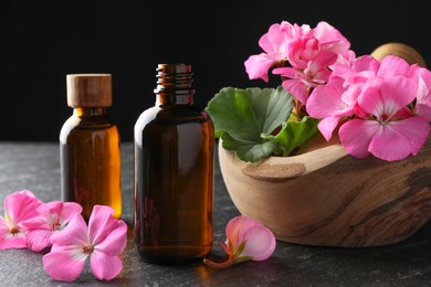 Photo of Bottles of geranium essential oil, beautiful flowers and mortar with pestle on black table, closeup