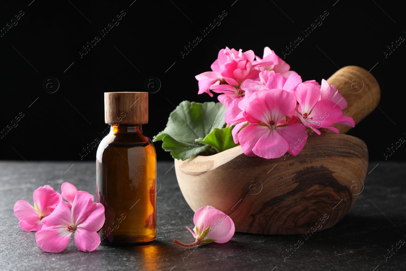 Photo of Bottle of geranium essential oil, beautiful flowers and mortar with pestle on black table, closeup