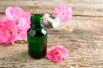 Photo of Geranium essential oil in bottle, pipette and beautiful flowers on wooden table, closeup. Space for text