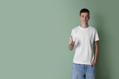 Teenage boy wearing white t-shirt and showing thumbs up on green background, space for text