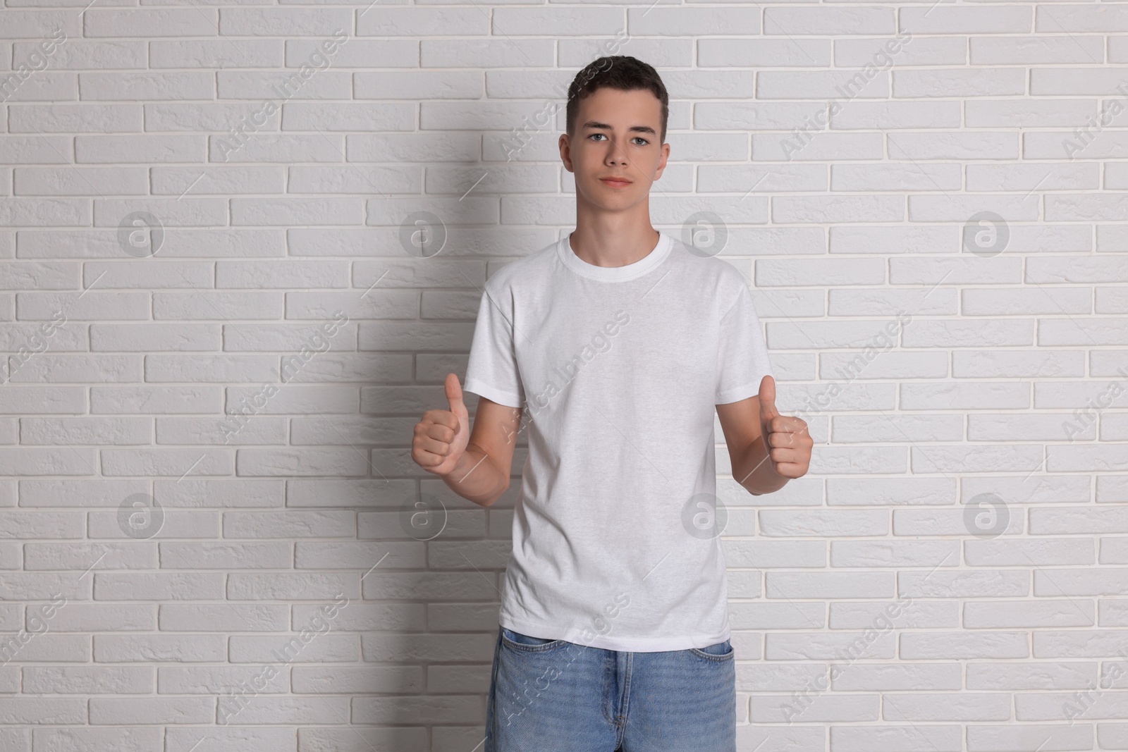 Photo of Teenage boy wearing t-shirt and showing thumbs up near white brick wall