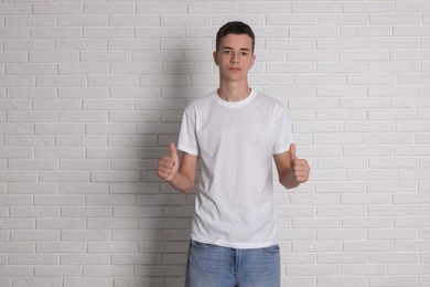 Photo of Teenage boy wearing t-shirt and showing thumbs up near white brick wall