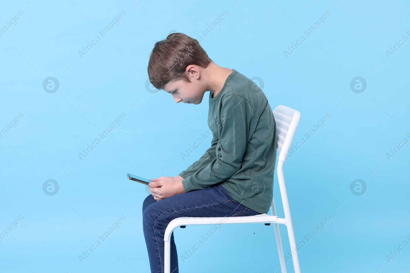 Photo of Boy with incorrect posture and phone sitting on chair against light blue background