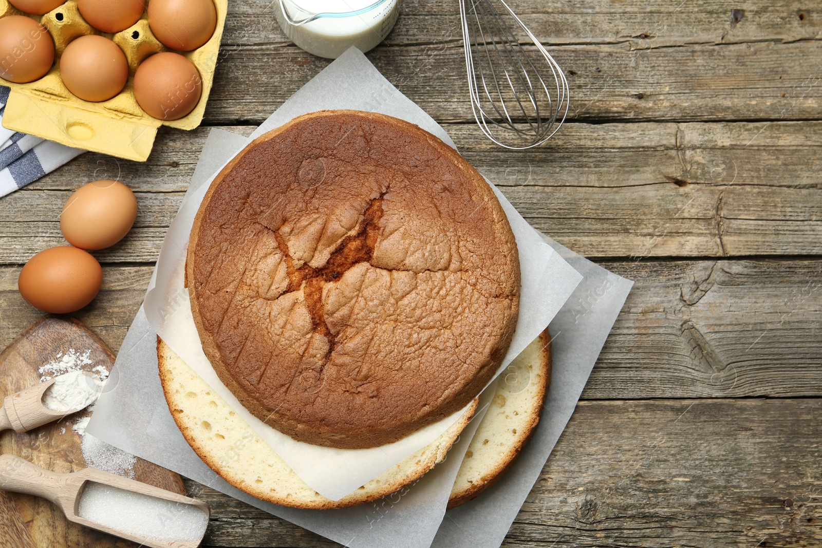 Photo of Delicious cut sponge cake, whisk and ingredients on wooden table, flat lay