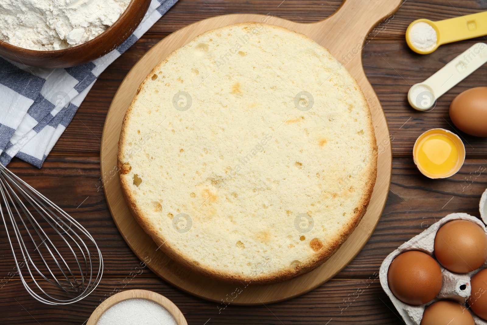 Photo of Delicious cut sponge cake, whisk and ingredients on wooden table, flat lay