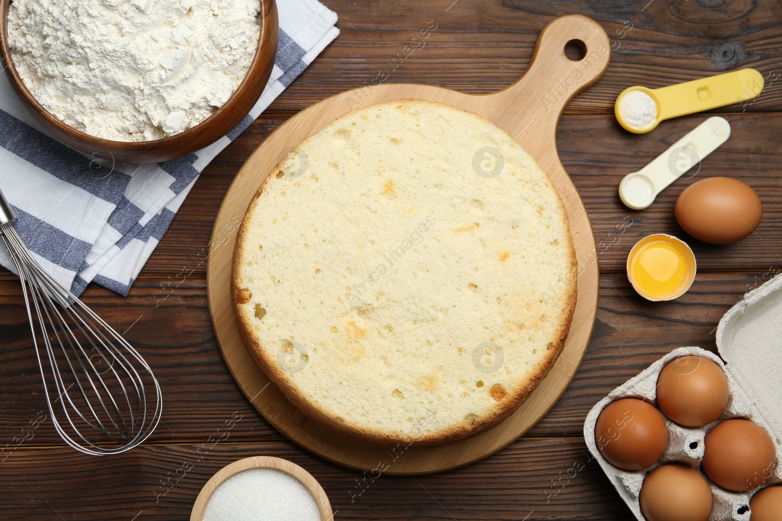 Photo of Delicious cut sponge cake, whisk and ingredients on wooden table, flat lay