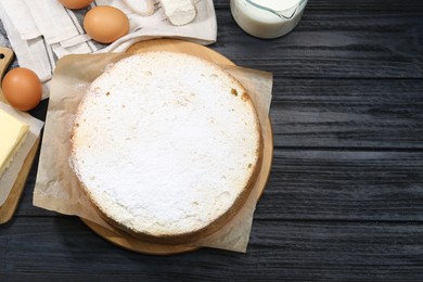 Photo of Delicious sponge cake with powdered sugar and ingredients on black wooden table, flat lay. Space for text