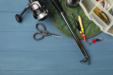 Fishing rod with spinning reel, baits, floats, scissors and net on blue wooden table, top view. Space for text