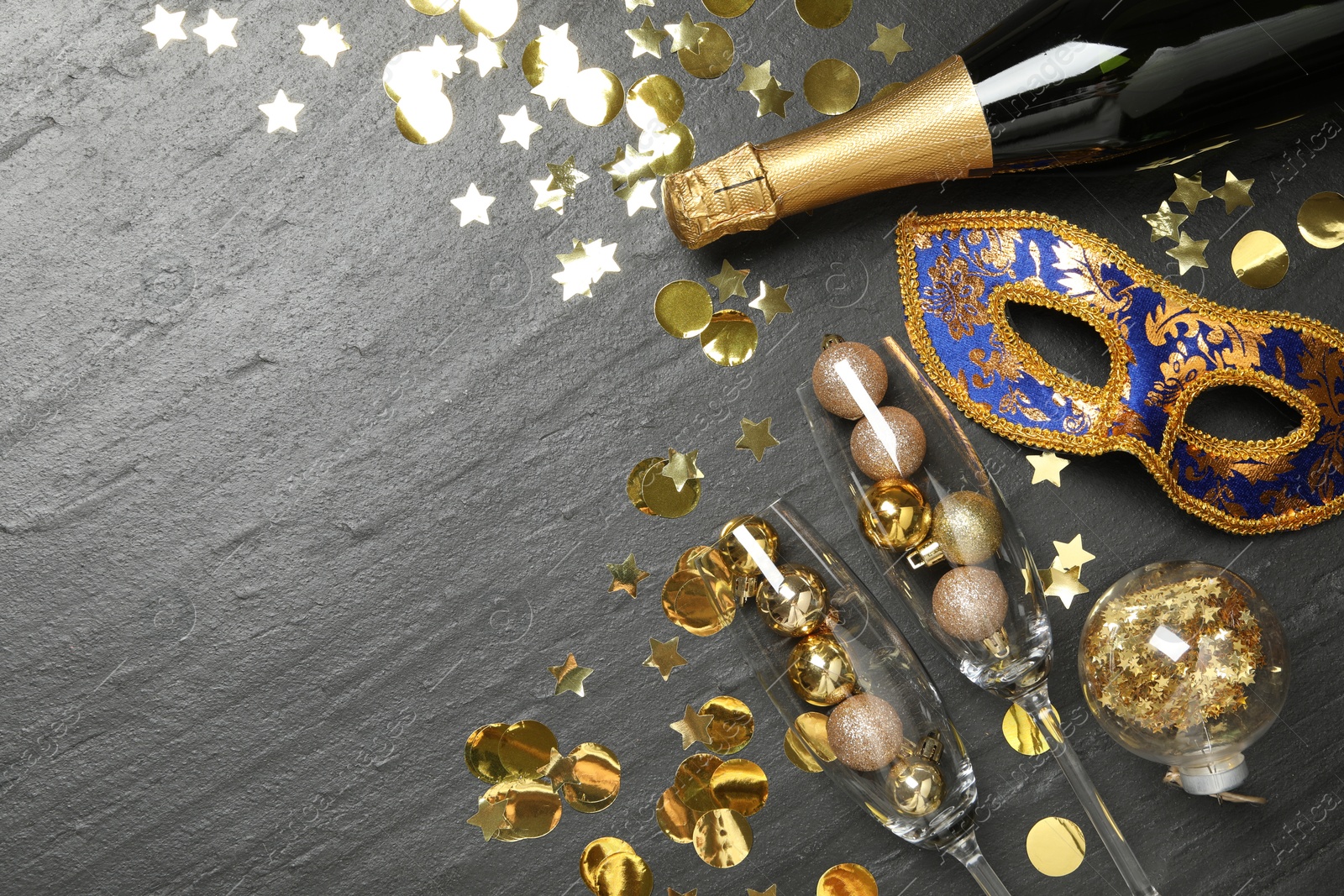 Photo of Beautiful golden carnival mask, bottle of sparkling wine, glasses with decorative balls and confetti on gray textured table, flat lay. Space for text