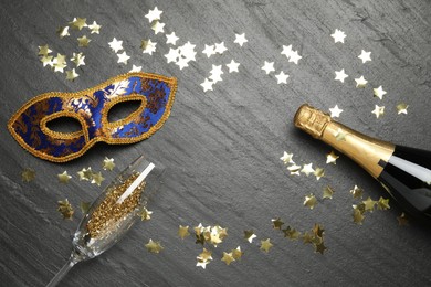 Beautiful golden carnival mask, bottle of sparkling wine, glass and confetti on gray textured table, flat lay. Space for text