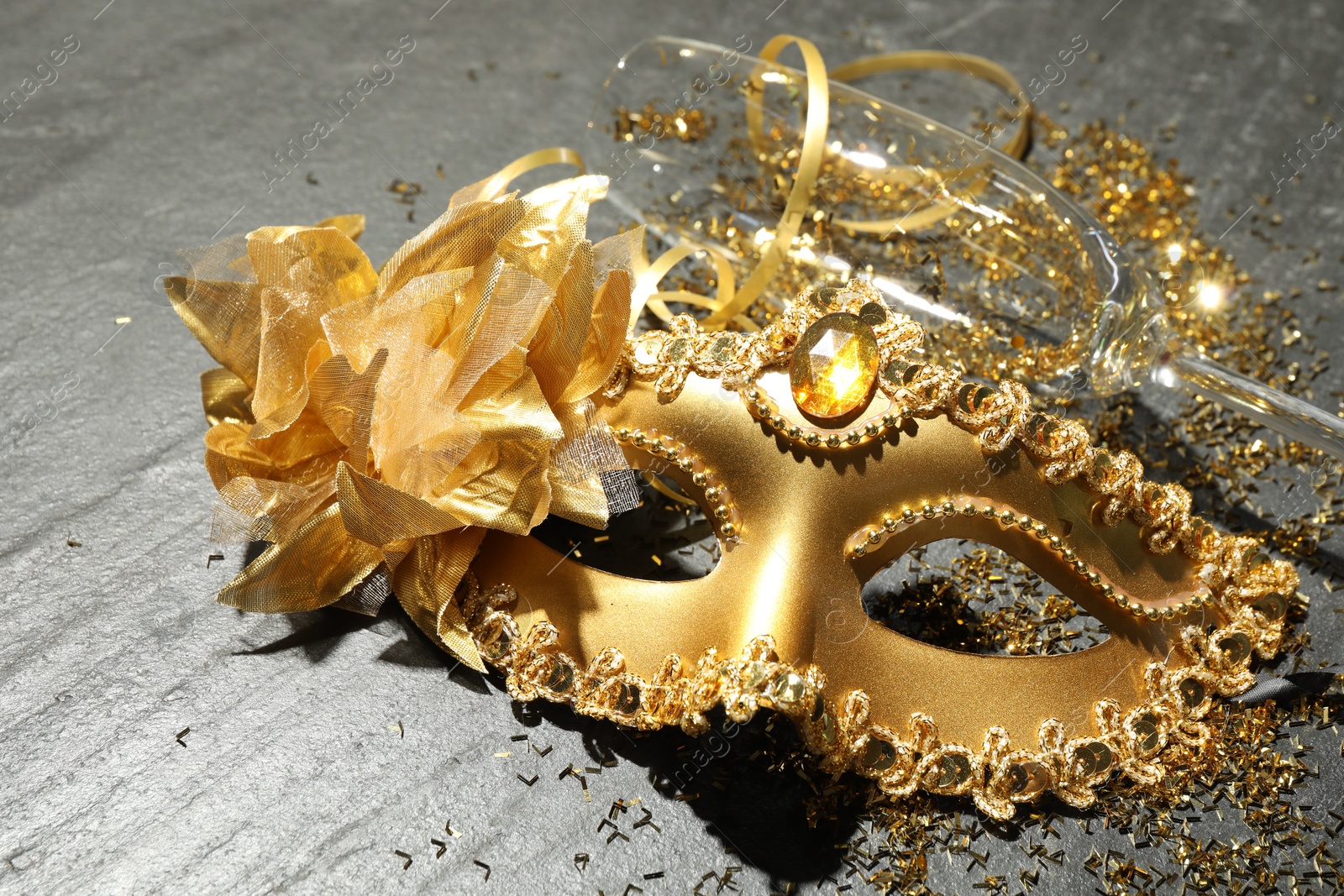 Photo of Beautiful golden carnival mask, glass and confetti on gray textured table, closeup