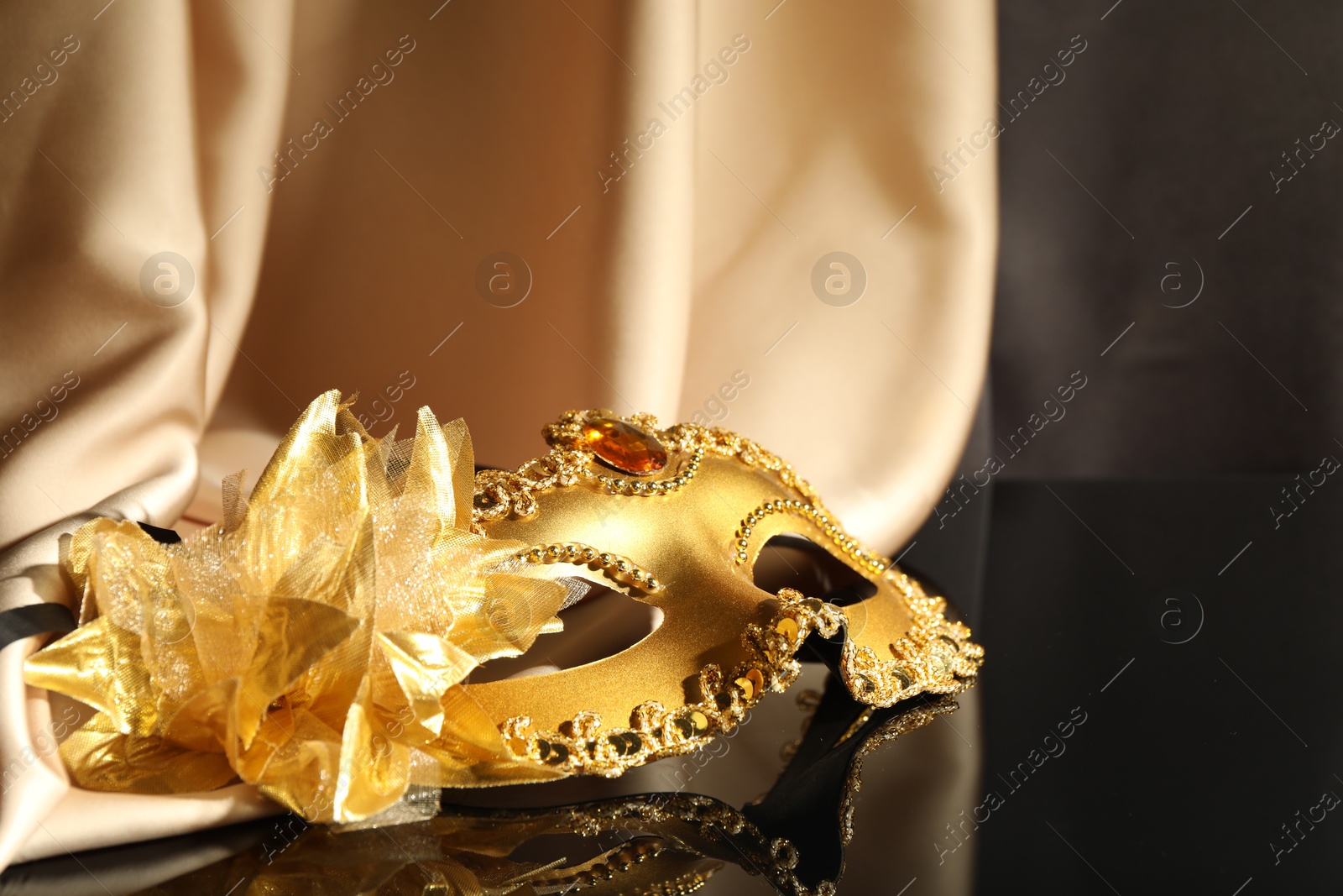 Photo of Beautiful golden carnival mask and beige fabric on black mirror surface