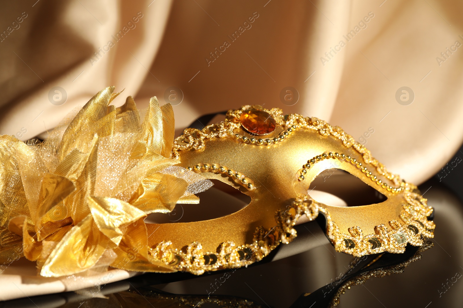 Photo of Beautiful golden carnival mask and beige fabric on black mirror surface, closeup