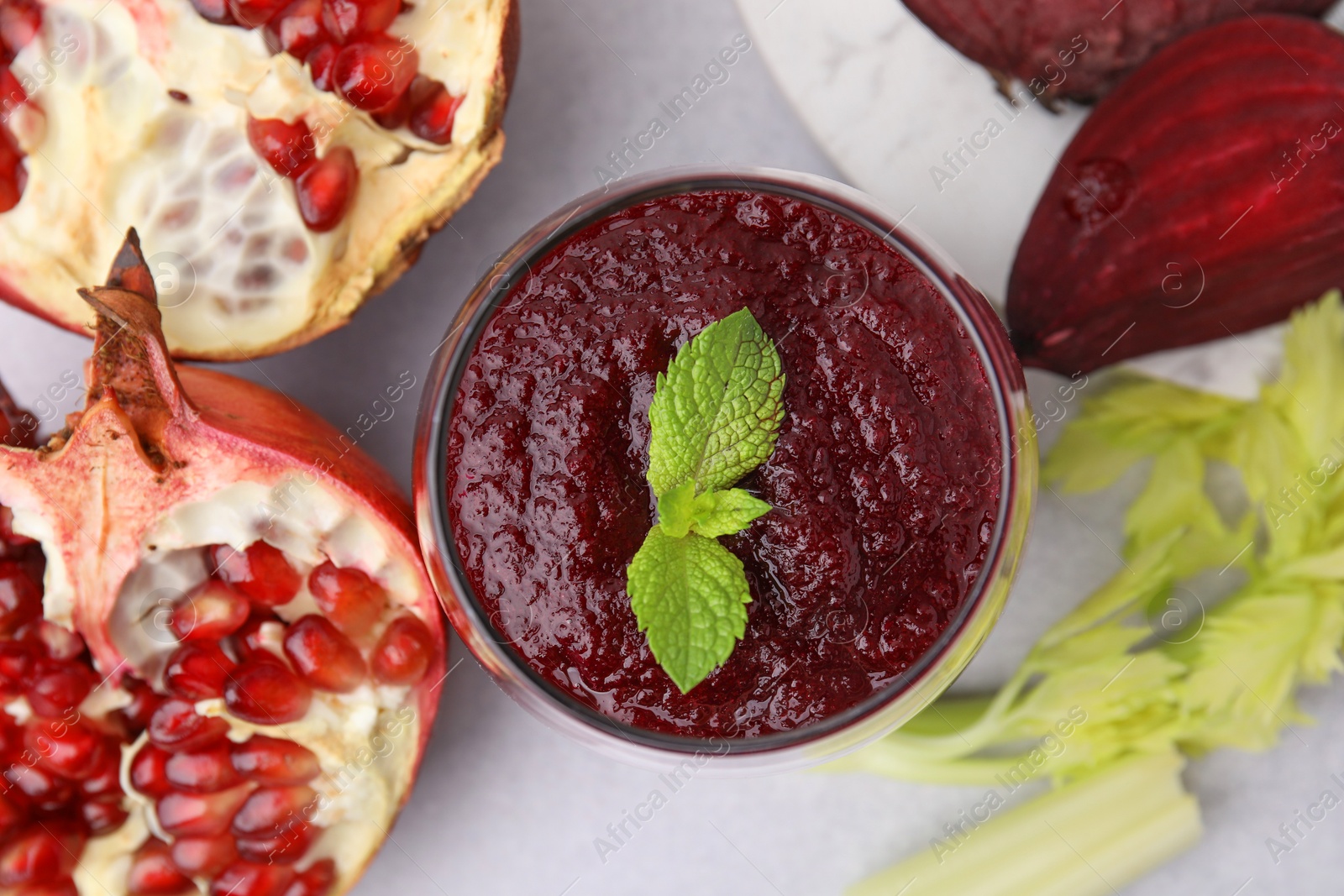 Photo of Tasty beetroot smoothie with mint in glass, fresh vegetables and pomegranate on light table, flat lay. Vegan drink