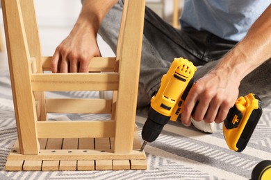 Photo of Man repairing wooden stool with electric screwdriver indoors, closeup